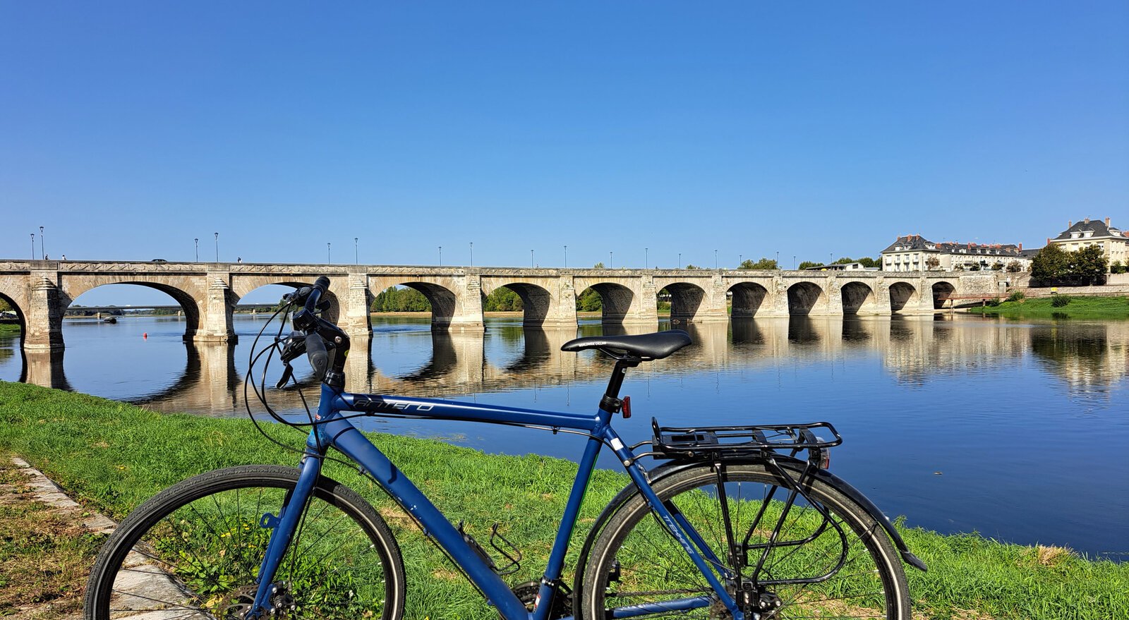 A blue bicycle with a rear rack stands on a grassy riverbank in front of a long stone arch bridge spanning a wide, calm river under a clear summer sky.