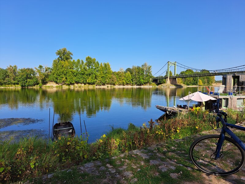 Un vélo appuyé au bord d'une rivière calme reflétant de grands arbres verdoyants, avec un pont suspendu visible en arrière-plan sous un ciel bleu d'été.