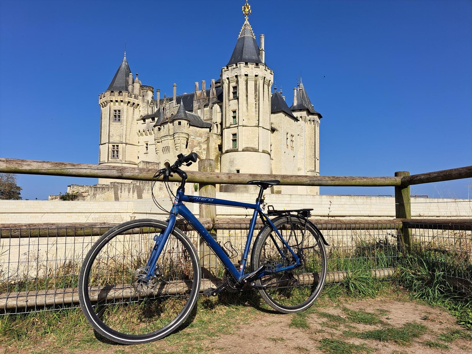 A blue touring bicycle leaned against a wooden fence in front of the grand turreted Château de Saumur under a clear blue sky.