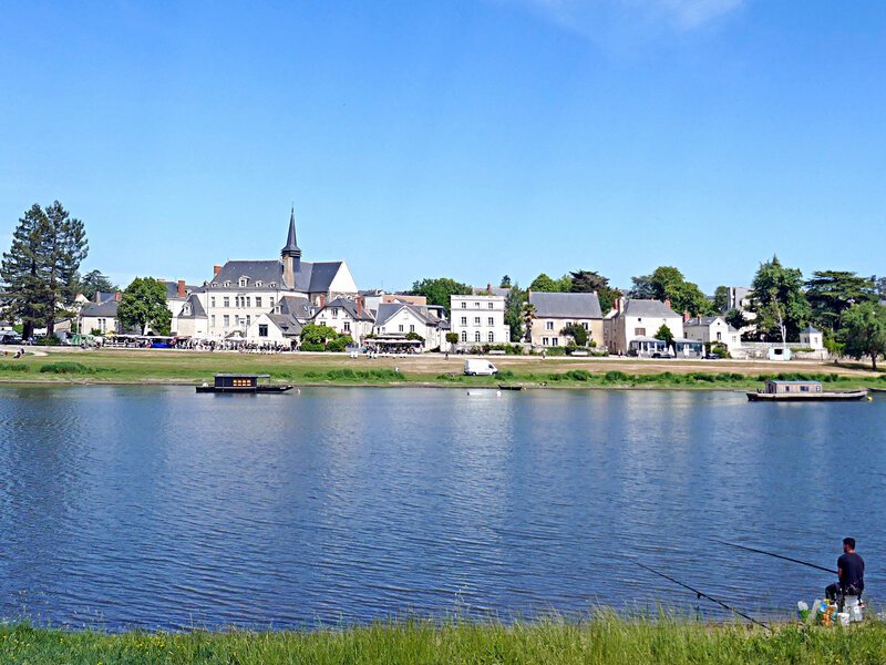 A person fishes from the grassy bank of a wide, calm river, looking across to the historic village of Candes-Saint-Martin with its stone church spire and white buildings under a clear blue summer sky.