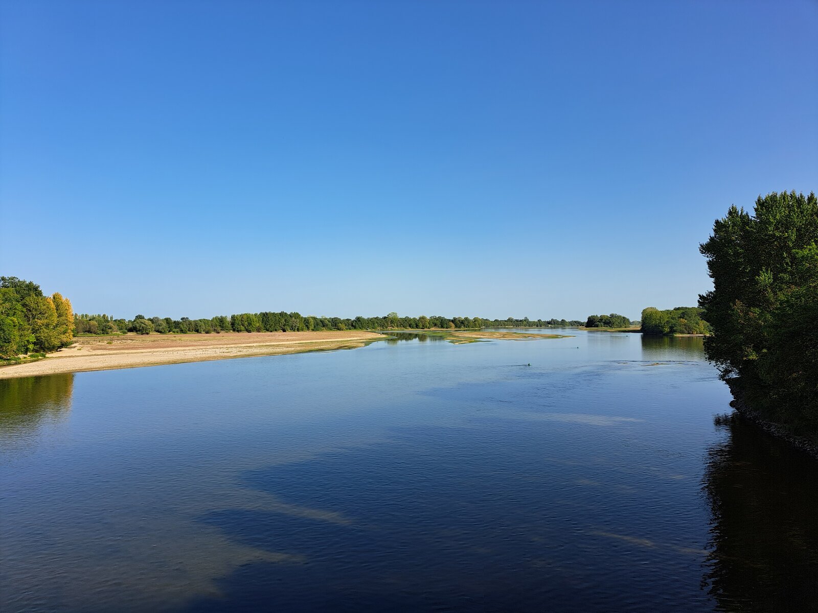 A wide, calm stretch of the Loire river under a clear blue summer sky, with sandy banks and green trees lining both shores.