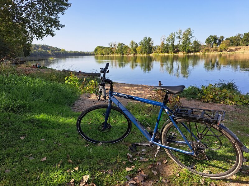 A blue touring bicycle parked on a grassy riverbank beside the calm, tree-lined Loire river on a sunny autumn day.