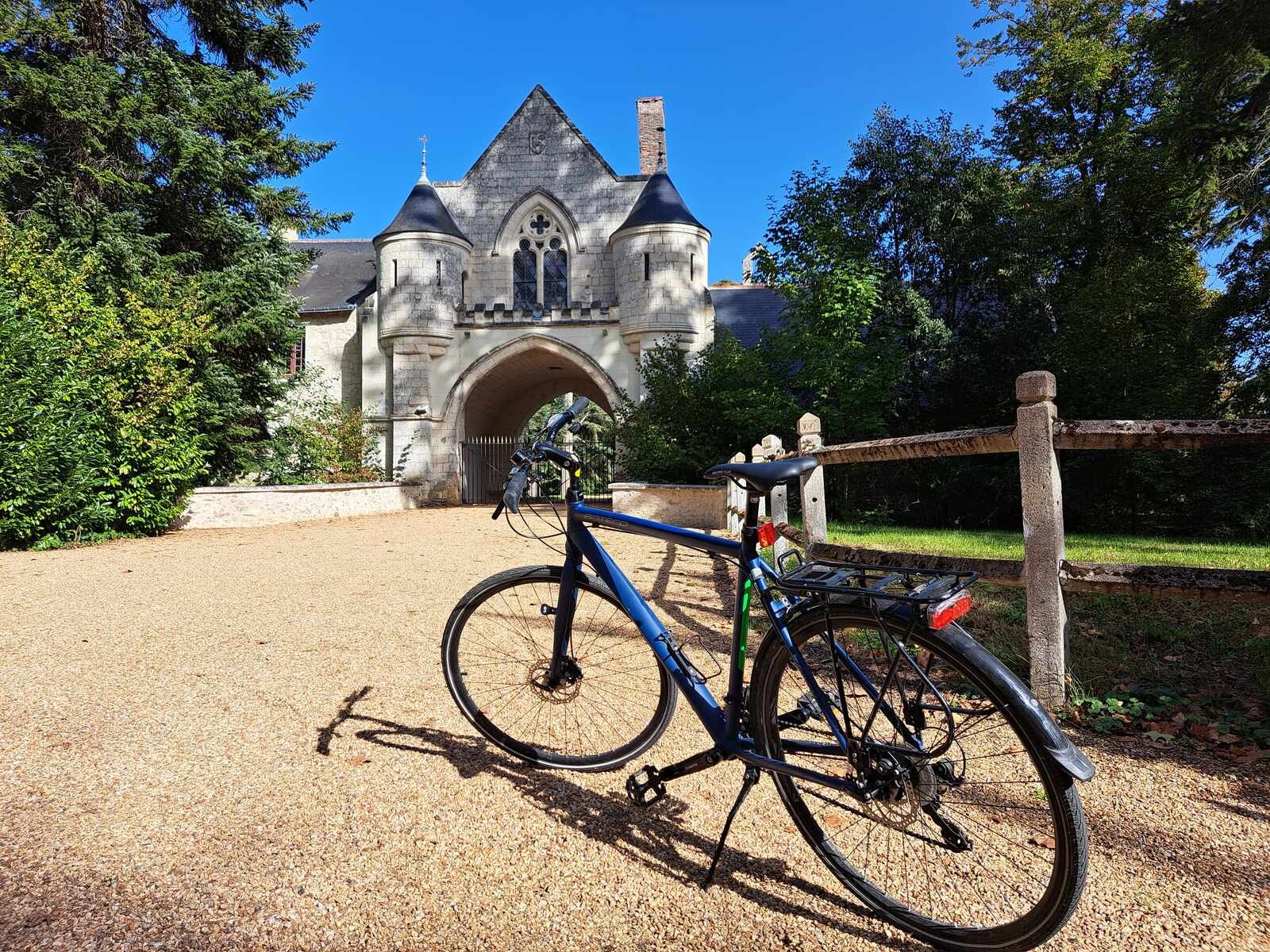 A blue bicycle leaning against a wooden fence post on a gravel path in front of a stone Gothic-arched gatehouse surrounded by mature trees on a sunny summer's day.