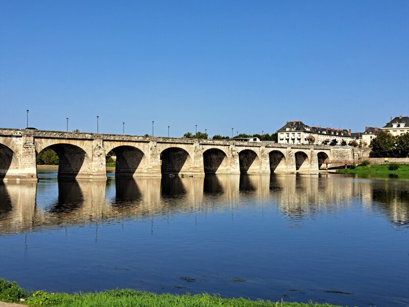 Un pont en pierre à arches multiples enjambant une rivière calme dans la vallée de la Loire, ses arches se reflétant nettement dans l'eau bleue et tranquille sous un ciel d'été sans nuages.