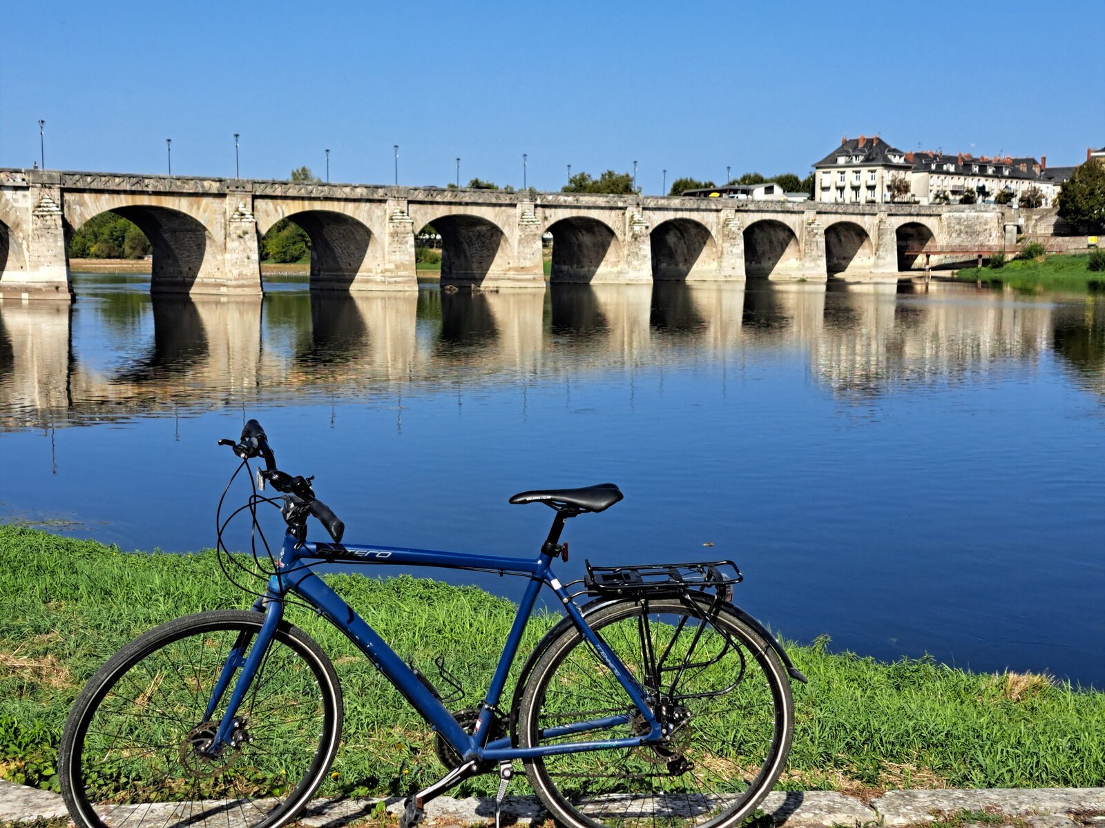 Un vélo de randonnée bleu garé au bord de la Loire, avec un pont en pierre à plusieurs arches et son reflet s'étirant sur les eaux calmes sous un ciel bleu dégagé.