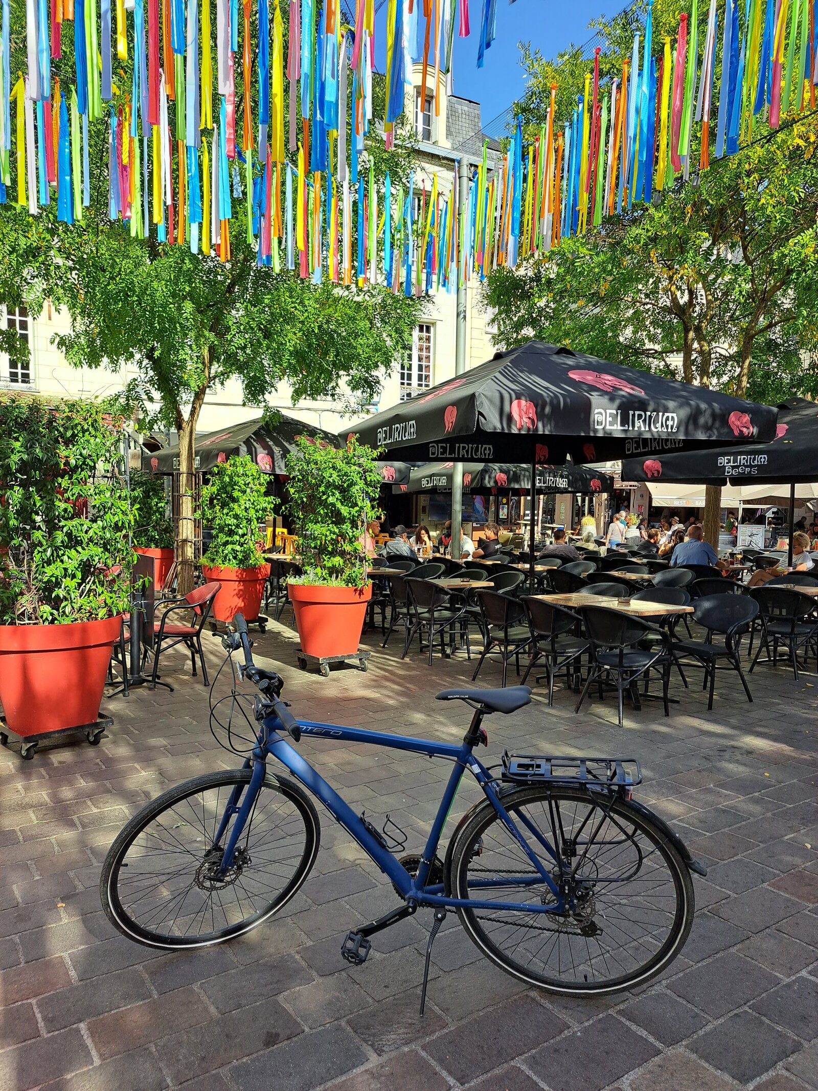 A blue touring bicycle parked on a sunny cobbled town square, with a bustling outdoor café terrace shaded by black umbrellas and colourful hanging ribbon decorations overhead.