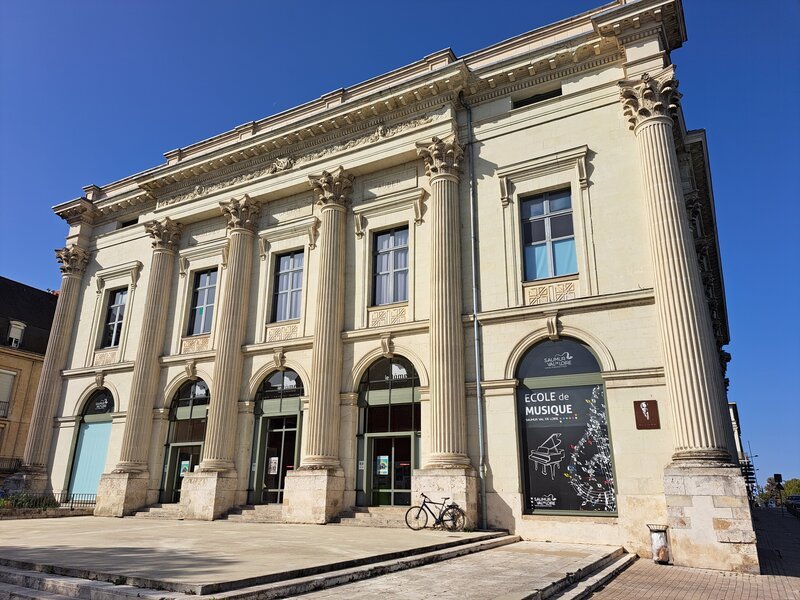 The neoclassical stone facade of La Cité de la Musique, featuring tall Corinthian columns and arched doorways beneath a clear blue sky, with a bicycle parked at the entrance steps.