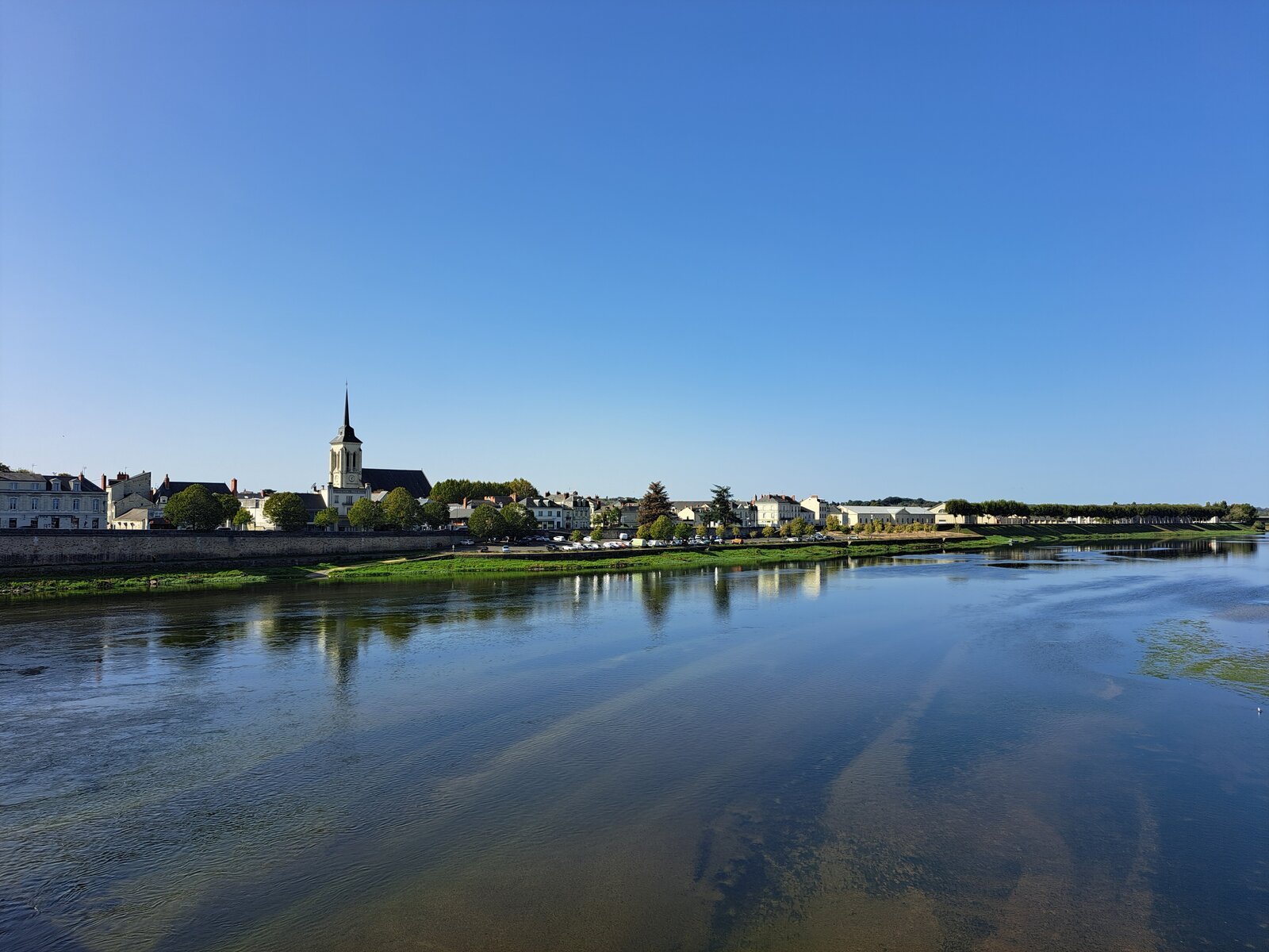 A wide, calm stretch of the Loire river reflects a clear blue sky, with a small French town featuring a church spire and low rooftops lining the far bank.