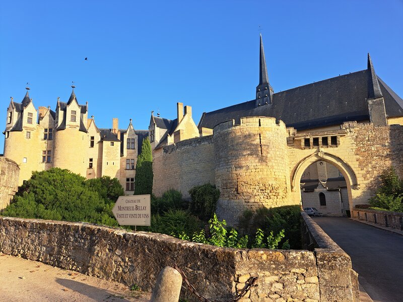 Château médiéval baigné de soleil aux murs de pierre claire, avec une flèche d'église gothique et une entrée en arc de cercle, vu depuis un vieux pont de pierre sous un ciel bleu limpide.