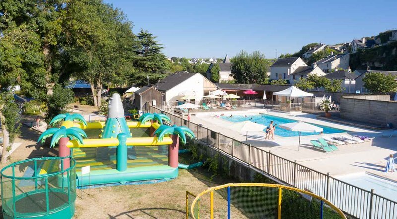 An aerial view of an outdoor swimming pool complex at Camping Isle Verte, featuring a large pool with swimmers, a colourful tropical-themed inflatable water play area with palm tree decorations, and sun umbrellas, surrounded by trees and residential buildings on a bright summer day.