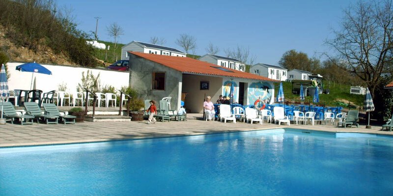 A large outdoor swimming pool at Camping Le Thouet, with sun loungers and parasols arranged around the terrace, and white holiday accommodation buildings set into a wooded hillside behind.