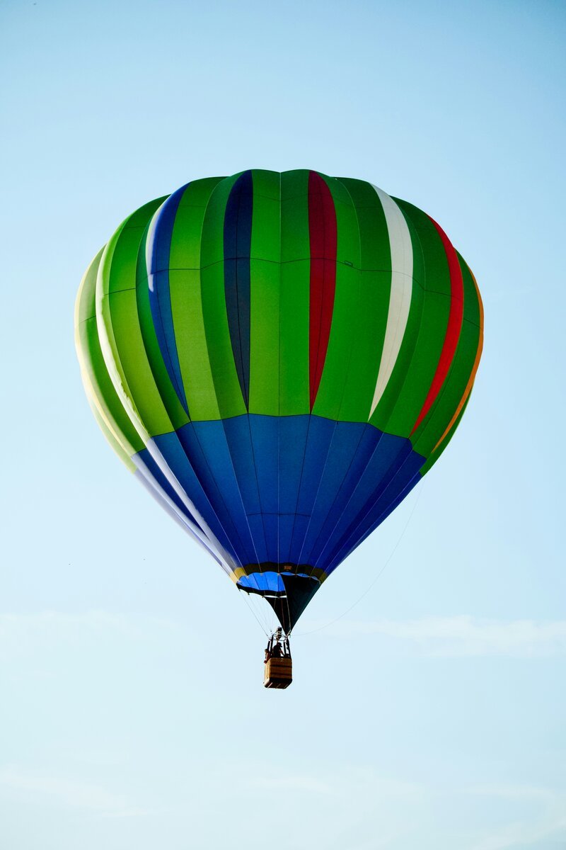A large hot air balloon with vivid green, blue, red and white vertical stripes floats against a pale blue sky, with a small passenger basket visible beneath.