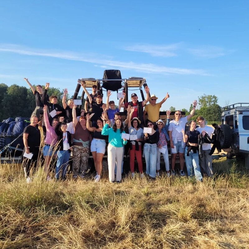 A large group of smiling people with arms raised in celebration standing in a golden field in front of a hot air balloon basket after landing.