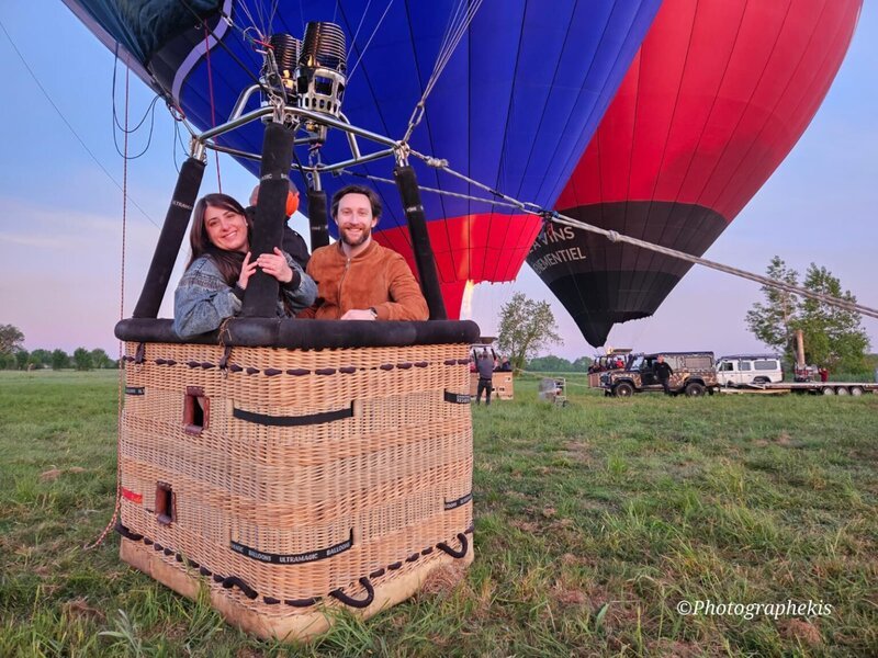 A smiling couple stand together in a wicker hot air balloon basket on the ground, with the colourful balloon inflating above them in a green field at dusk or dawn.