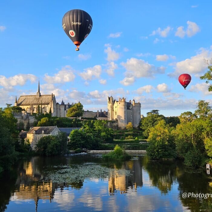 Three hot air balloons float above the medieval château and church of Montreuil-Bellay on a sunny summer evening, their reflections visible in the calm river below.