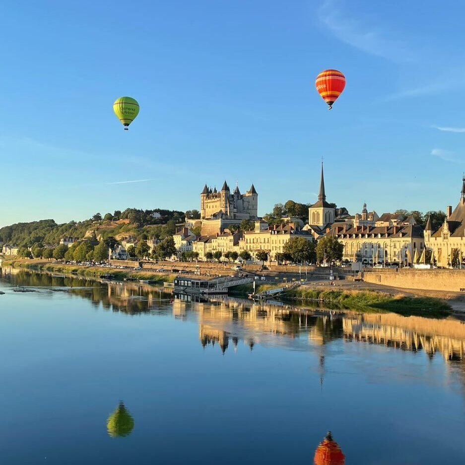 Two colourful hot air balloons float above the historic town of Saumur on the Loire, with the turreted château and church spire reflected in the calm river below on a clear summer morning.