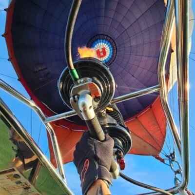 A view looking up from inside a hot air balloon basket, showing a gloved hand operating the burner with an open flame heating the purple and red envelope above against a clear blue sky.
