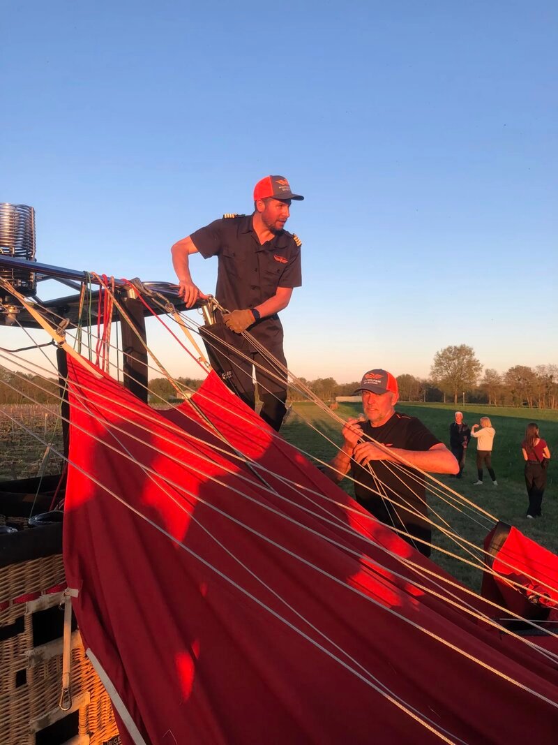 Two crew members in matching dark shirts and red caps work together to manage the rigging ropes of a red hot air balloon as it deflates on landing in an open field at dusk or dawn.