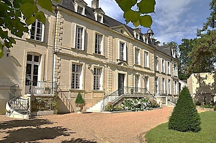 The elegant stone façade of Château de Beaulieu, a classic French manor house with symmetrical windows, wrought-iron balustrades, a double staircase, and neatly kept gardens on a sunny day.