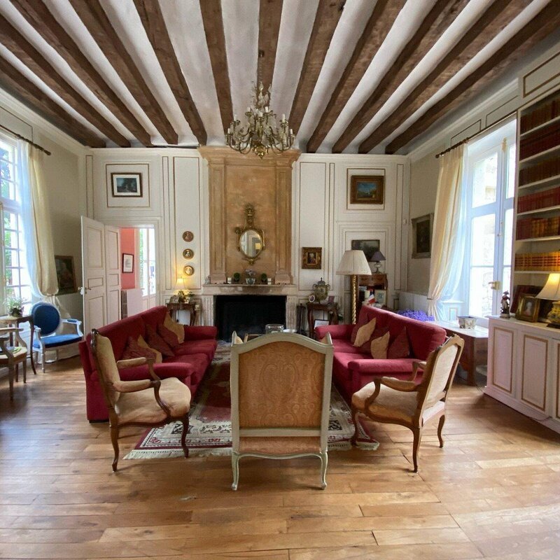 A grand salon at Château de Beaulieu featuring exposed timber beam ceilings, a crystal chandelier, red velvet sofas and gilded armchairs arranged around a large stone fireplace, with polished parquet floors and tall windows letting in natural light.