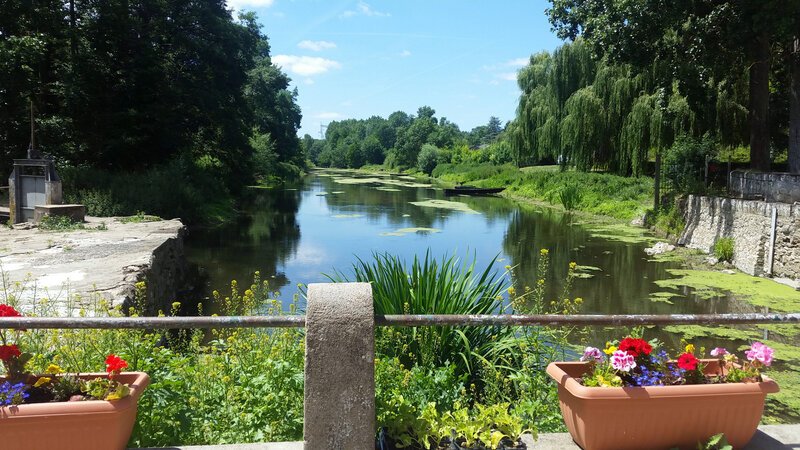 Une rivière paisible vue depuis la rambarde d'une terrasse bordée de fleurs, encadrée d'arbres verdoyants sous un ciel bleu aux nuages légers, avec des fleurs en pot colorées au premier plan.