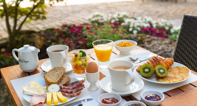 Un généreux petit-déjeuner continental servi sur un plateau à une table en plein air, comprenant un œuf à la coque, un croissant, du pain tranché, du bacon, du kiwi frais et une salade de fruits, un jus d'orange, un café, de la confiture et une petite omelette, avec des jardins fleuris visibles en arrière-plan.