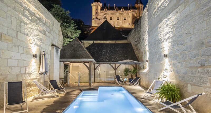 Une petite piscine extérieure illuminée avec des chaises longues, nichée dans une cour aux murs de pierre, de nuit, avec un château médiéval visible en arrière-plan.
