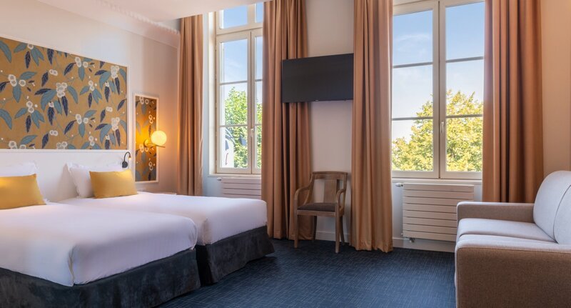 Une chambre d'hôtel lumineuse et élégante avec deux lits simples habillés de linge blanc, des rideaux dorés encadrant de hautes fenêtres, une télévision murale, un panneau mural à motifs floraux et un canapé gris au premier plan.