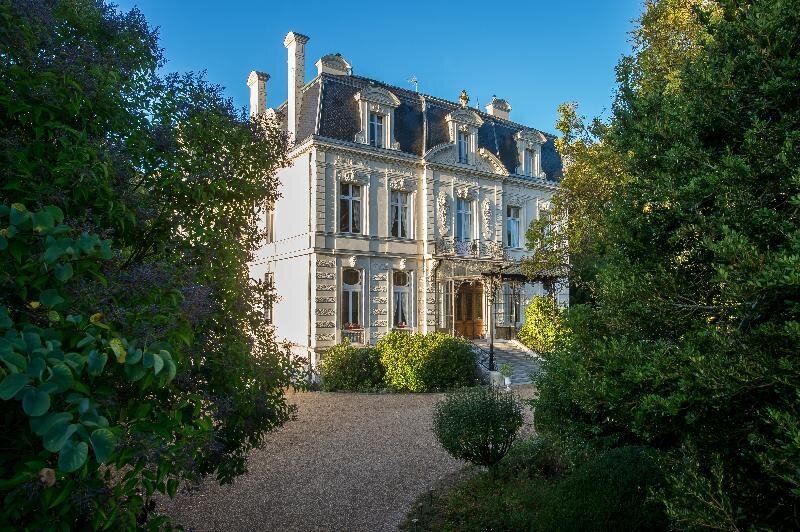 A grand white French château with a mansard roof and ornate stone façade, surrounded by mature trees and lush gardens with a gravel driveway leading to the entrance.