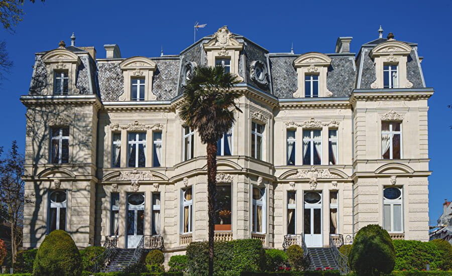 A grand French château with a mansard roof, ornate stone facade, tall shuttered windows, and a palm tree in the foreground, photographed on a clear sunny day.