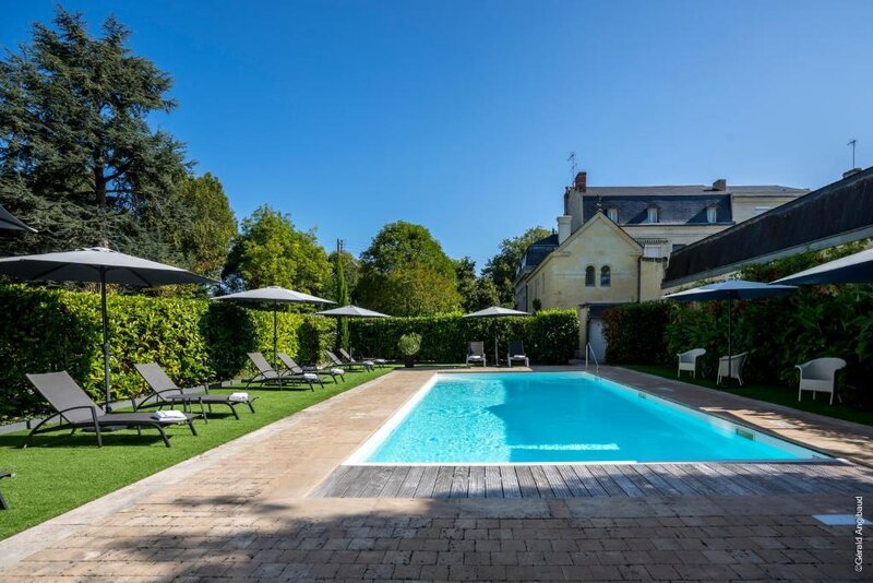 A rectangular outdoor swimming pool surrounded by sun loungers and white parasols, set within a manicured garden beside a traditional French stone property on a clear summer's day.