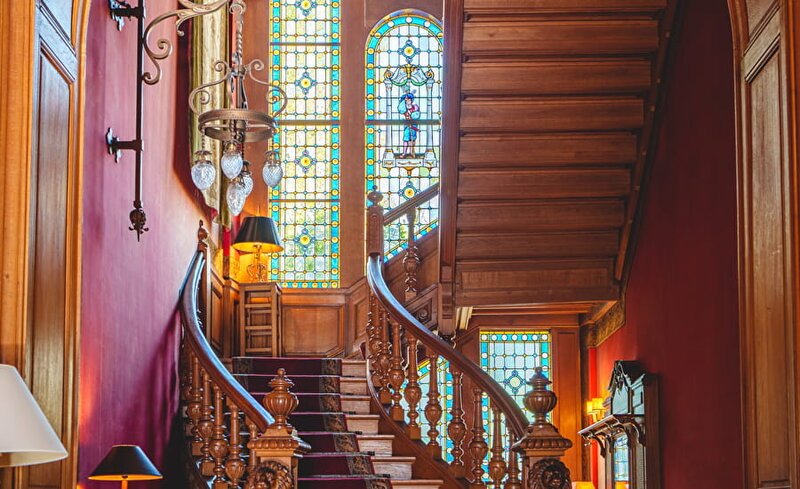 An ornate carved wooden spiral staircase sweeps upward inside a grand Loire Valley château, flanked by richly panelled red walls, a tall arched stained-glass window, and antique wall lanterns.