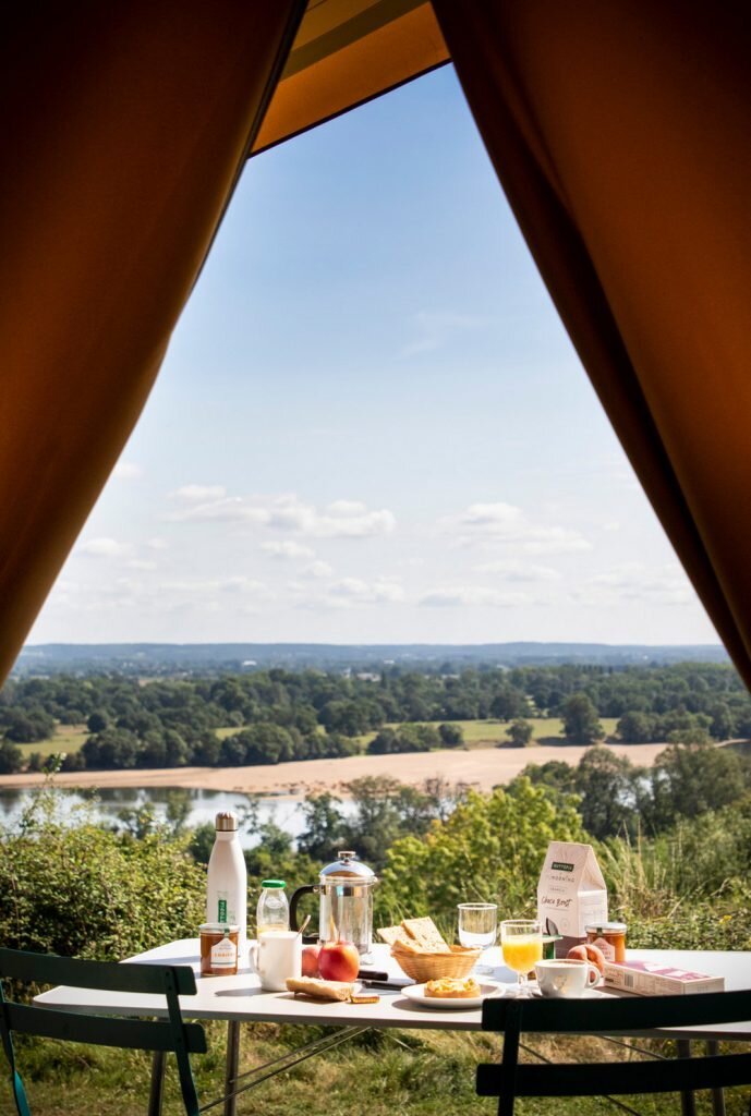 Une table de petit-déjeuner dressée avec jus d'orange, café, pain et fruits, vue depuis l'ouverture triangulaire d'une tente en toile, avec un panorama grandiose sur la vallée de la Loire et ses collines boisées sous un ciel bleu d'été.