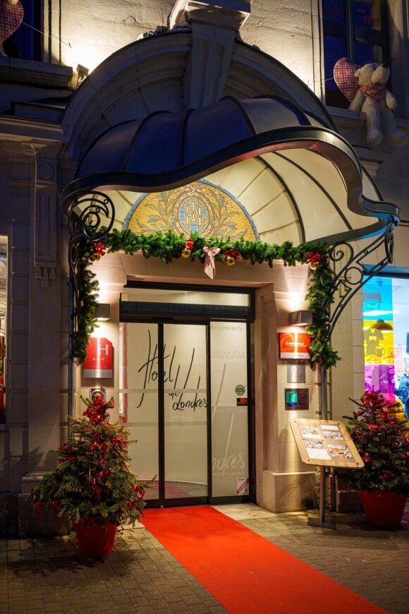 L'entrée illuminée de l'Hôtel Le Londres de nuit, ornée de guirlandes de Noël et de baies rouges au-dessus de portes vitrées automatiques, avec un tapis rouge menant à l'intérieur sous un élégant auvent en dôme bleu.