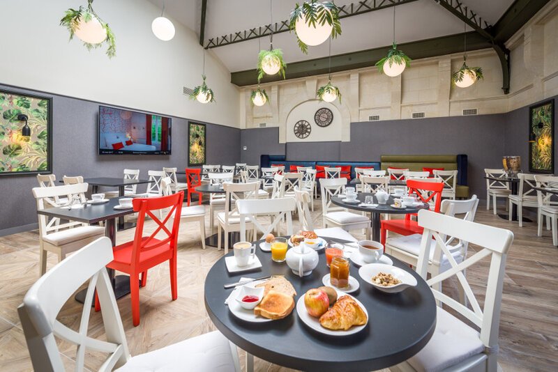 A bright, modern hotel dining room with round black tables set for breakfast, including croissants, orange juice and jam, surrounded by white and red chairs, hanging globe lights with trailing plants, and artwork on grey walls.
