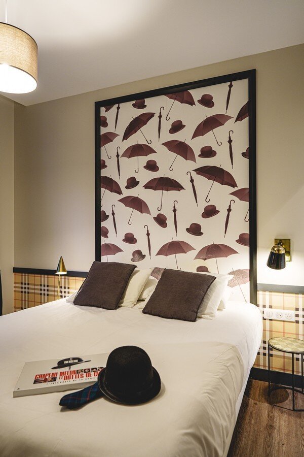 A stylishly decorated hotel double bed with a large framed wallpaper panel featuring scattered red umbrellas and bowler hats above the headboard, with a bowler hat and magazine resting on the white bedding.