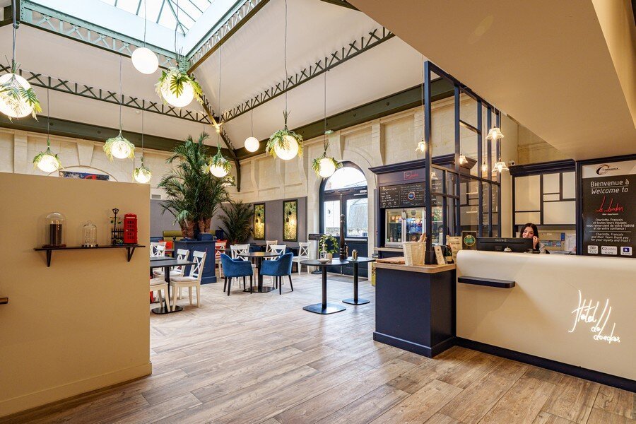 A bright, modern restaurant interior with wooden flooring, pendant globe lights, blue upholstered chairs, a reception counter, and a glass skylight roof.