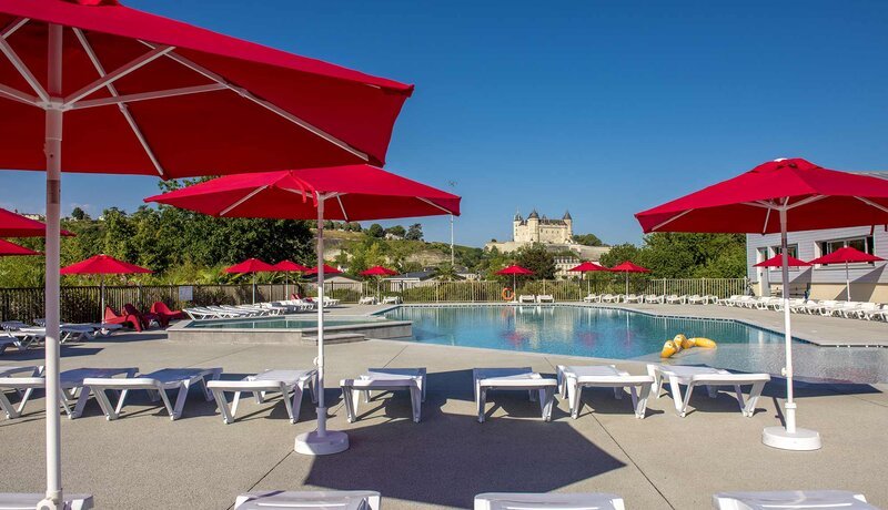 A sunny resort swimming pool terrace with rows of white sun loungers shaded by large red parasols, with a château and green hills visible in the background.