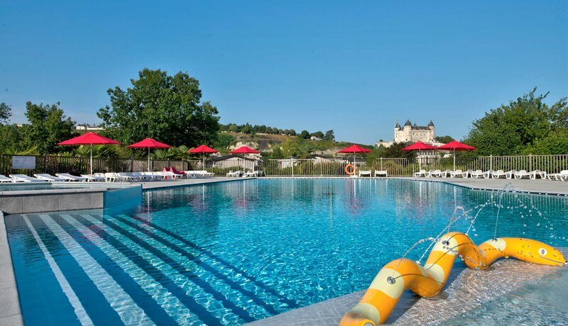 Une grande piscine extérieure aux eaux bleu cristal, avec des bouées gonflables jaunes au premier plan, des parasols rouges et des transats le long du bord de la piscine, et un château perché sur une colline visible entre les arbres en arrière-plan, sous un ciel d'été d'un bleu éclatant.