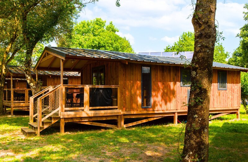 A raised timber lodge with a covered wooden deck and ramp, surrounded by trees on a green summer campsite.