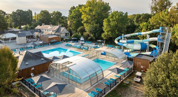 Aerial view of La Brèche campsite's outdoor pool complex featuring a large swimming pool, a covered pool section, blue sun loungers, and a turquoise water slide surrounded by trees.
