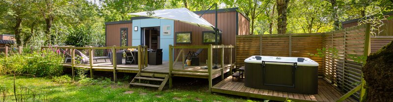 A geodesic dome glamping lodge with a wraparound timber deck and an outdoor hot tub, set amongst leafy trees in a woodland garden.
