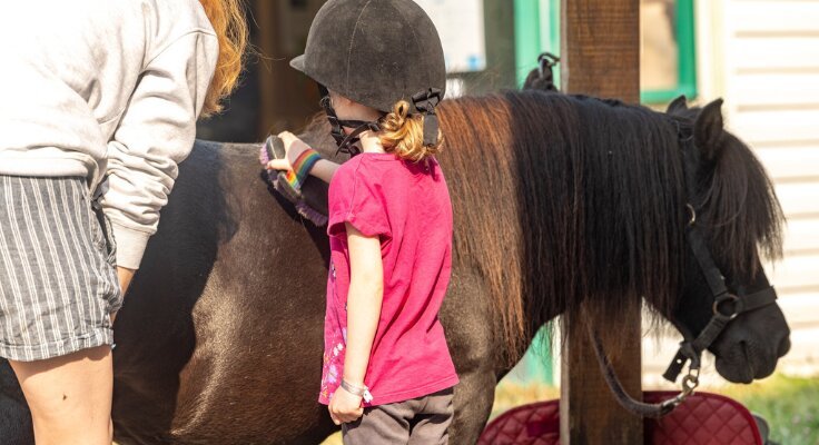 A young girl in a pink top and riding helmet sits on a dark brown pony beside a stable, with an adult helper standing close by.