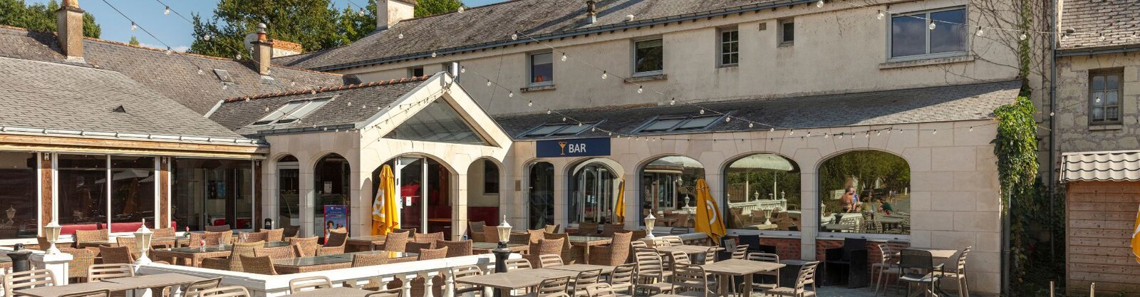 The exterior of La Brèche restaurant, showing a stone building with white arched colonnades, outdoor dining tables and chairs arranged on a terrace, and lush greenery surrounding the façade.