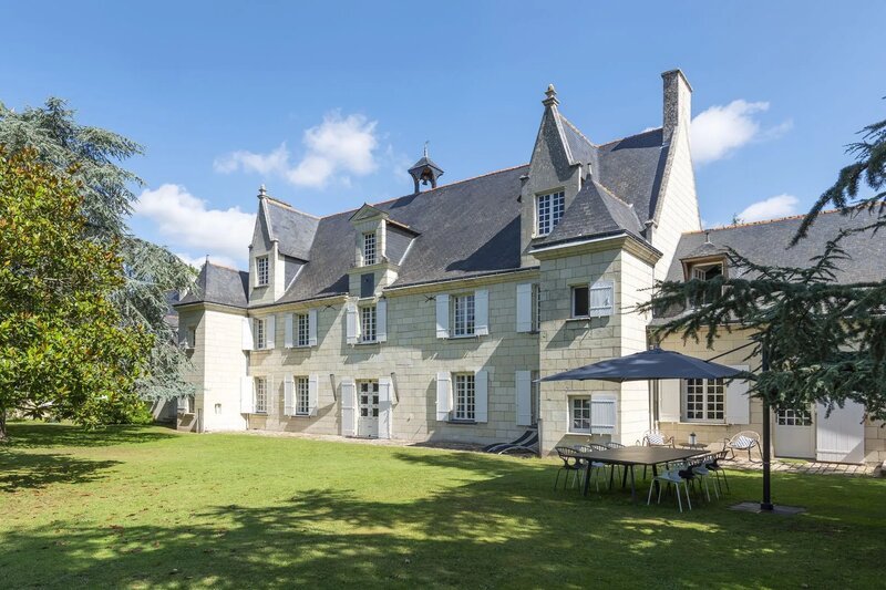 A grand French château with grey slate roofs and pale stone façade, set in a sweeping green lawn with mature trees and an outdoor dining table with a parasol in the foreground.