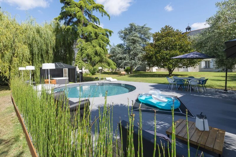 Une piscine extérieure ronde entourée d'une terrasse pavée avec des transats et un parasol bleu, nichée dans un jardin verdoyant avec de grands saules et chênes, par une belle journée d'été ensoleillée.