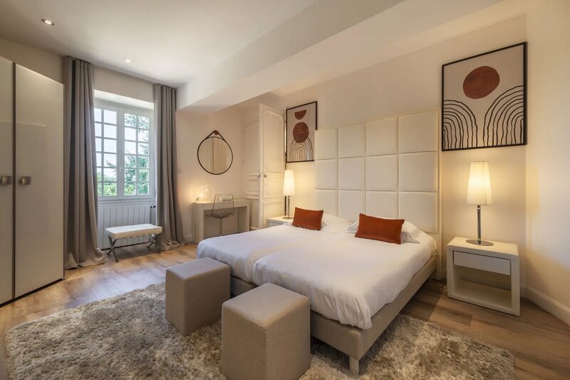 Une chambre twin lumineuse et élégamment décorée dans un château français, avec des panneaux muraux tapissés, des coussins d'accent en terracotta, un tapis moelleux, des lampes de chevet et une grande fenêtre donnant sur la verdure.