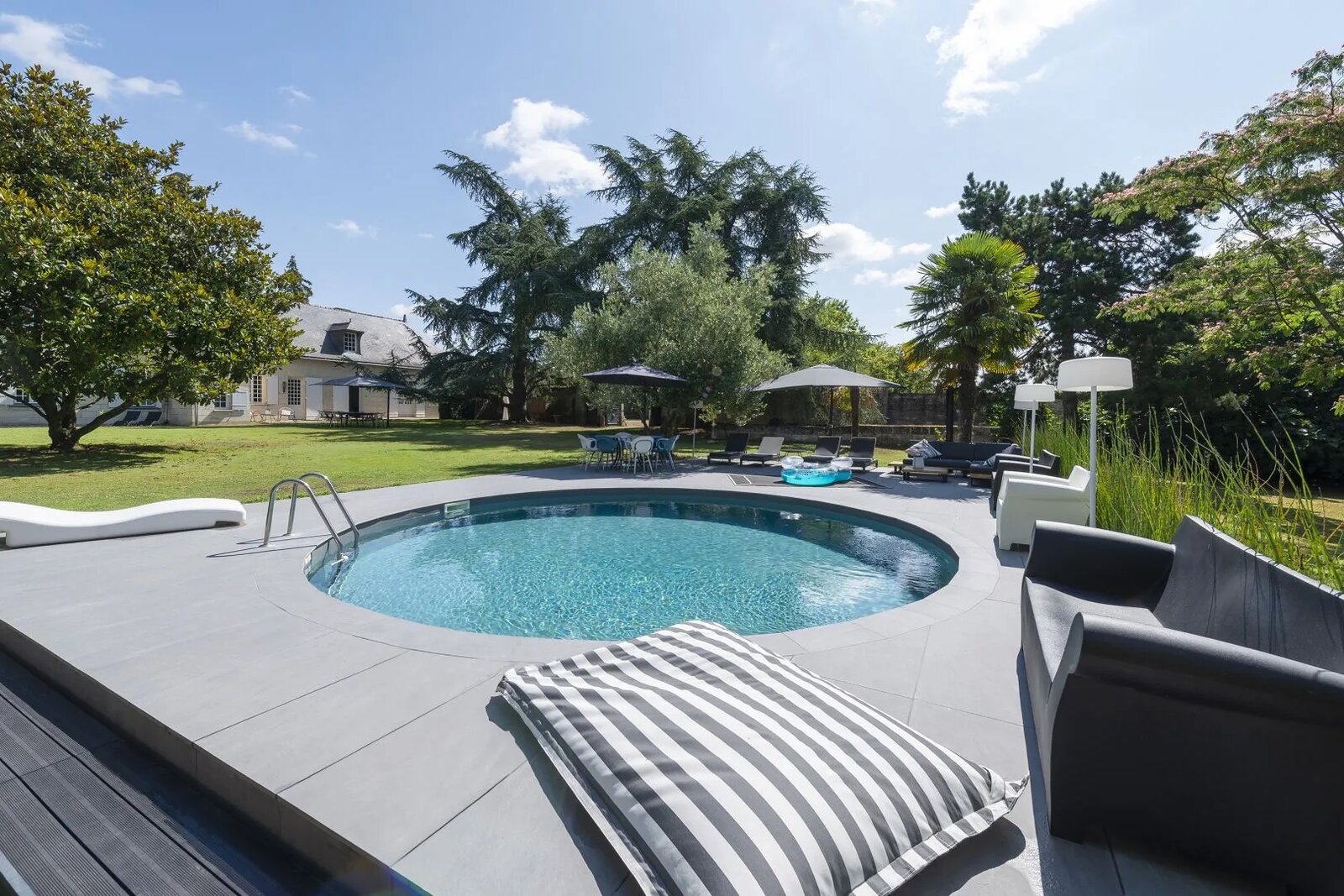 Une piscine extérieure circulaire encastrée dans une terrasse en pierre grise, entourée de chaises longues et d'arbres luxuriants par une belle journée d'été.